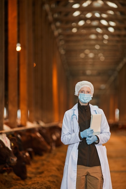 Vertikales Hüftporträt des weiblichen Tierarztes, der Maske am Bauernhof trägt und Kamera betrachtet, während Tablette hält