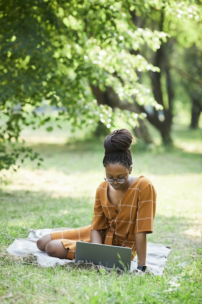Vertikales Porträt der afroamerikanischen jungen Frau mit Laptop im Park beim Sitzen auf Gras, Kopierraum