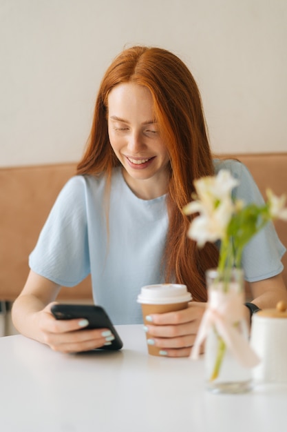 Vertikales Porträt der lächelnden jungen Frau, die Smartphone-Nachricht schreibt und heißen Kaffee trinkt, der am Schreibtisch im gemütlichen Café sitzt. Kaukasische Dame der hübschen Rothaarigen, die Freizeitbeschäftigung im Café hat.