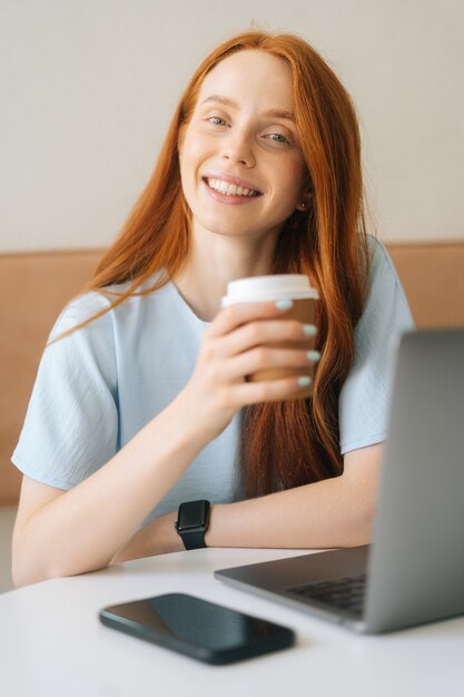 Vertikales Porträt der Nahaufnahme der glücklichen jungen schönen Frau, die Tasse mit heißem Kaffee hält, der am Schreibtisch mit Laptop im Café sitzt und Kamera betrachtet. Hübsche kaukasische Dame der Rothaarigen entfernt, die arbeitet oder studiert.