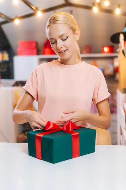 Vertikales Porträt des Designers der jungen Frau, der Weihnachtsgeschenkbox einwickelt, die rotes Band bindet und