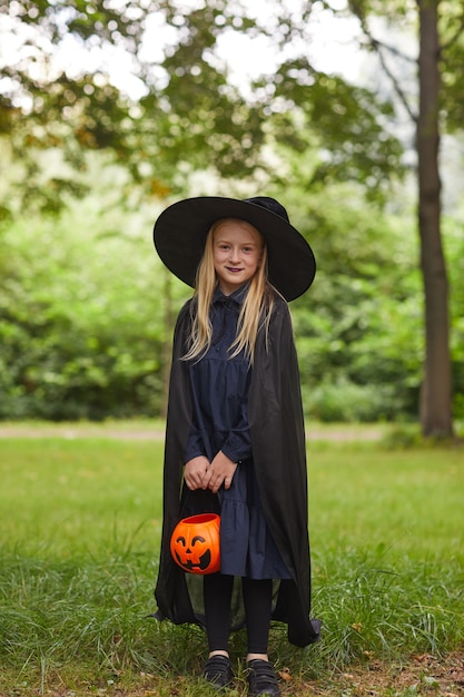 Vertikales Porträt des lächelnden Teenager-Mädchens in voller Länge, gekleidet als Hexe, die draußen aufwirft und Halloween-Eimer hält