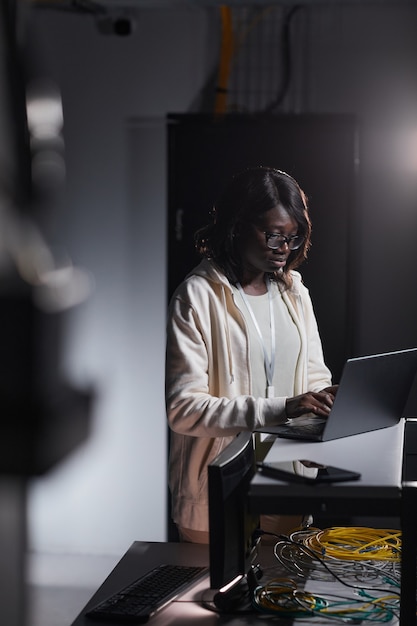 Vertikales Porträt einer afroamerikanischen Netzwerkingenieurin, die einen Laptop verwendet, während sie im dunklen Serverraum arbeitet