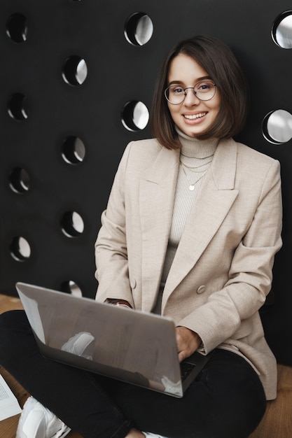 Vertikales Porträt einer eleganten, gutaussehenden Frau mit Brille und Blazer, die mit Laptop auf dem Boden sitzt und Hausaufgaben studiert, Projektschreibbericht vorbereitet und die Kamera glücklich lächelt