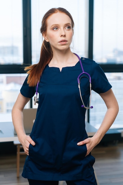 Vertikales Porträt einer ernsthaften rothaarigen Ärztin in medizinischer Uniform, die auf dem Hintergrund des Fensters wegschaut