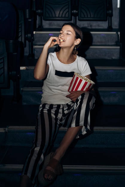Vertikales Porträt einer Frau, die in einem Kino auf der Treppe sitzt und eine Popcorn-Eimer-Schüssel hält
