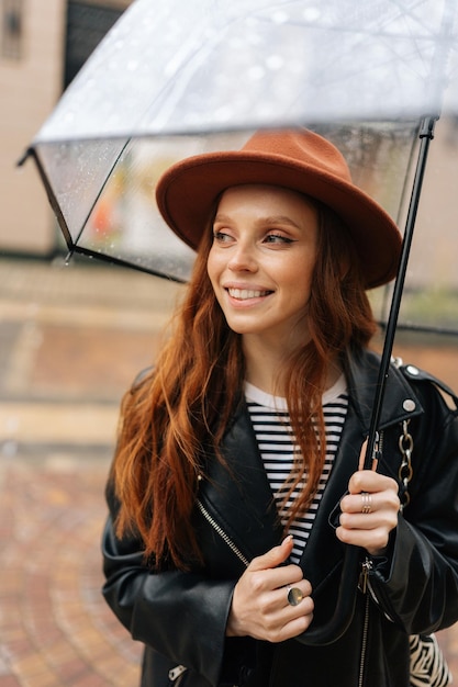 Vertikales Porträt einer fröhlichen, ingwerfarbenen jungen Frau mit elegantem Hut, die mit transparentem Regenschirm auf der Stadtstraße steht und Regenwetter im Freien genießt