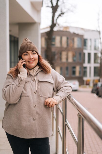 Vertikales Porträt einer fröhlichen übergewichtigen Frau mit warmem Hut und Jacke, die an einem bewölkten Herbsttag auf einem Smartphone in der Nähe eines Geländers an der Stadtstraße steht