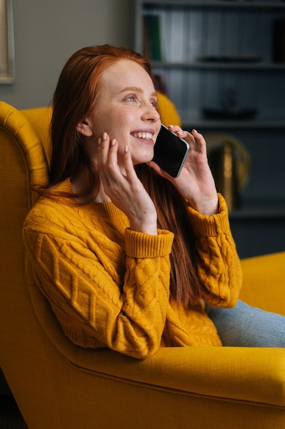 Vertikales Porträt einer glücklich lächelnden jungen Frau, die auf dem Smartphone spricht und zu Hause in einem gelben Stuhl sitzt Hübsche Dame, die ein angenehmes Gespräch hat, das per Telefon spricht