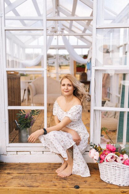 Vertikales Porträt einer glücklichen blonden jungen Frau in einem wunderschönen Kleid, die an der Tür des Sommers sitzt
