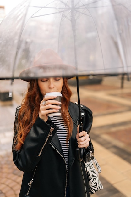 Vertikales Porträt einer glücklichen jungen Frau mit modischem Hut, die mit transparentem Regenschirm im kalten Regen der Stadt steht und heißen Kaffee trinkt