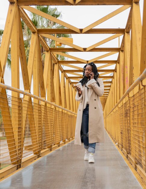 Vertikales Porträt einer jungen afroamerikanischen Frau mit Zöpfen, die mit einer Tasse Kaffee auf einer gelben Brücke spaziert und mit ihrem Handy spricht