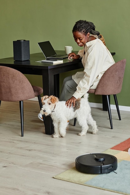 Vertikales Porträt einer jungen schwarzen Frau, die Hund streichelt, während sie von zu Hause aus in einer modernen Wohnung arbeitet