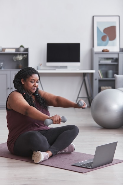 Vertikales Porträt einer kurvigen Afroamerikanerin, die zu Hause mit Hanteln trainiert, während sie auf einer Yogamatte sitzt und Online-Trainingsvideos ansieht, Platz kopieren