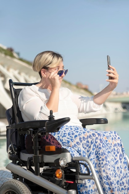 Vertikales Porträt einer lächelnden Frau mit Behinderung im Rollstuhl, die ein Selfie-Foto mit Blick hinter eine Sonnenbrille macht