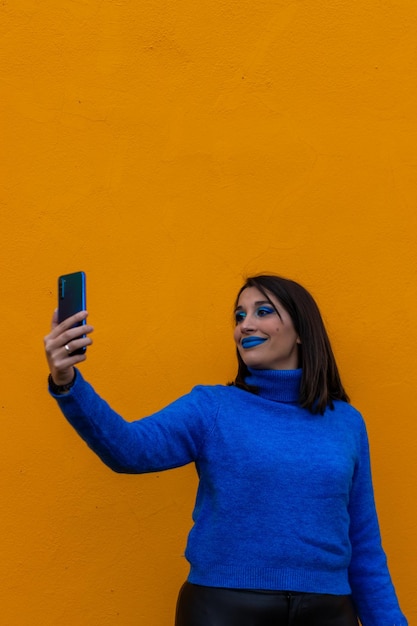 Vertikales Porträt einer lächelnden Frau mit Make-up und blauer Kleidung, die ein Selfie mit ihrem Handy auf gelbem Hintergrund macht