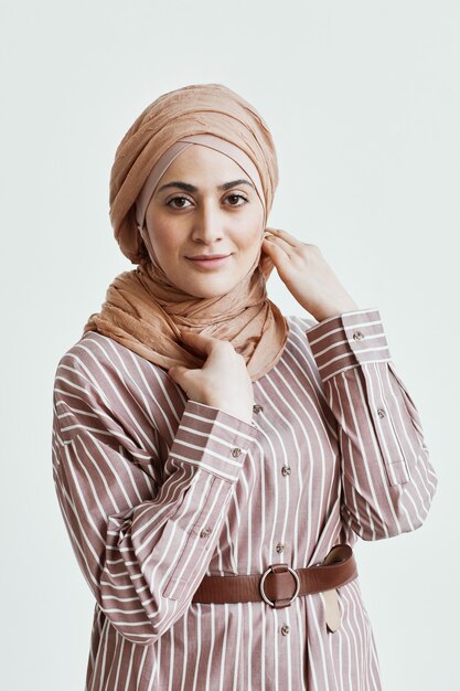Vertikales Porträt einer modernen Frau aus dem Nahen Osten, die in die Kamera schaut und ein Kopftuch trägt, während sie gegen eine weiße Wand steht