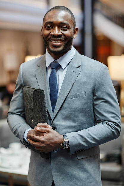 Vertikales Porträt eines erfolgreichen afroamerikanischen Geschäftsmannes, der in die Kamera lächelt, während er in der Hotellobby steht