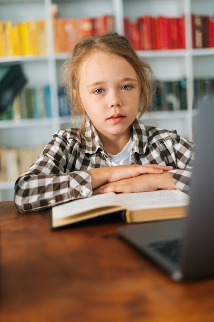 Vertikales Porträt eines fokussierten Grundschulmädchens, das mit Laptop und Papierbuch am Tisch sitzt