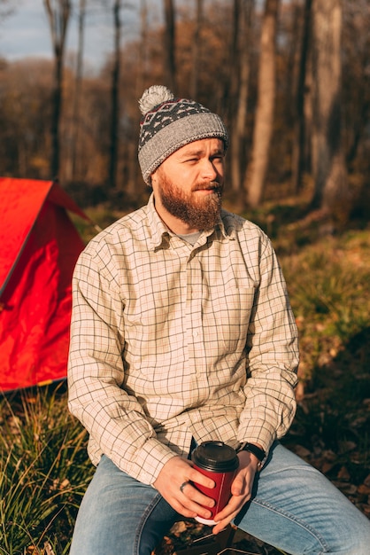 Vertikales Porträt eines jungen bärtigen Hipsters sitzt vor seinem Zelt im Wald.