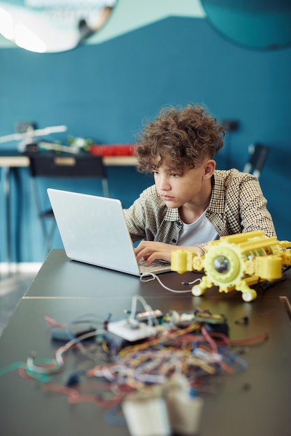 Vertikales Porträt eines jungen lockigen Jungen mit Laptop in der Schule während des Ingenieurunterrichts und des Programms