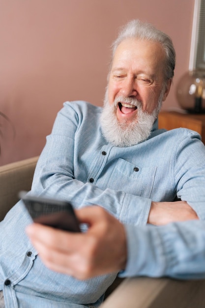 Vertikales Porträt eines lachenden, reifen Mannes, der sein Smartphone mit der mobilen Online-App in der Hand hält und fröhlich auf den Bildschirm schaut, während er auf einem Sessel sitzt. Bärtiger älterer Mann, der nach seiner Pensionierung zu Hause soziale Medien genießt