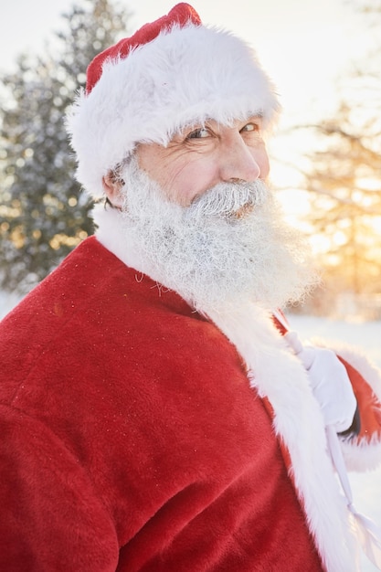 Vertikales Porträt eines lächelnden Weihnachtsmanns, der draußen im Winterwald in die Kamera schaut und sa trägt