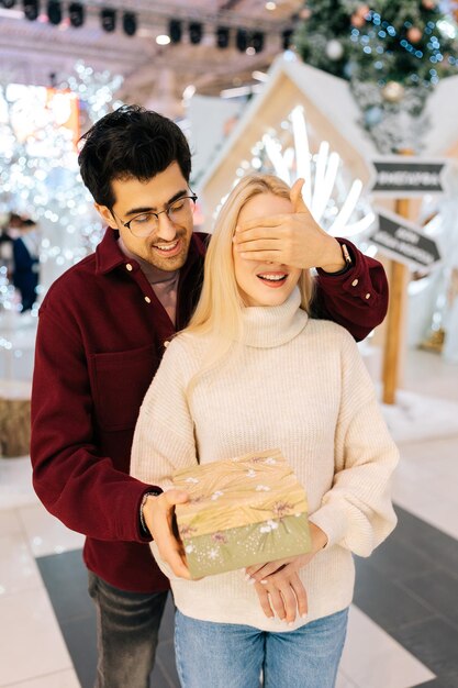 Vertikales Porträt eines liebevollen jungen Mannes, der die Augen bedeckt, glücklich lächelnde Freundinnen, die in der Halle des Einkaufszentrums am Weihnachtsabend stehen