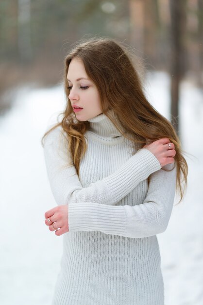 Vertikales Porträt eines schönen jungen Mädchens in einem weißen Pullover gegen den Winterwald