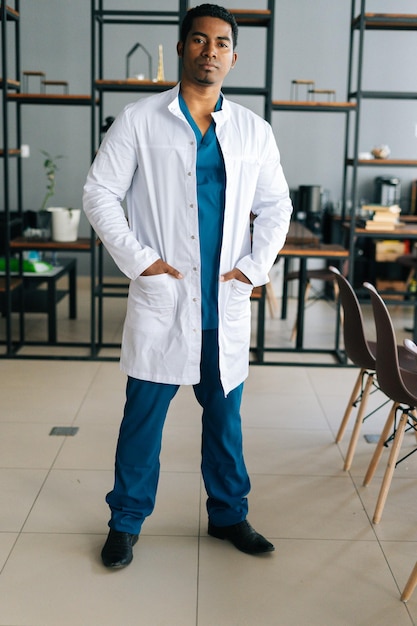 Vertikales Porträt eines selbstbewussten afroamerikanischen männlichen Arztes in medizinischer Laboruniform, der im Krankenhausbüro steht und in die Kamera blickt. Selbstbewusster Praktiker, der am Arbeitsplatz posiert.