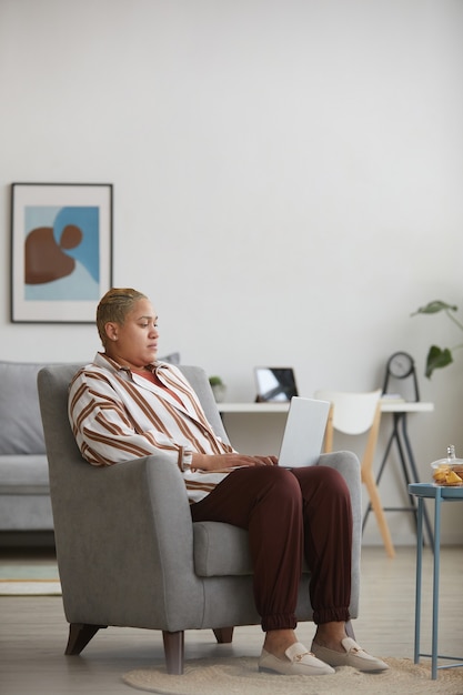 Vertikales Porträt in voller Länge einer modernen Mixed-Race-Frau mit Laptop, während sie zu Hause im Sessel sitzt, Kopienraum