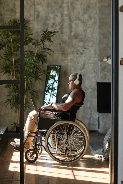 Vertikales Porträt in voller Länge einer zeitgenössischen tätowierten Frau mit Behinderung, die Musik hört, während sie sich zu Hause entspannt