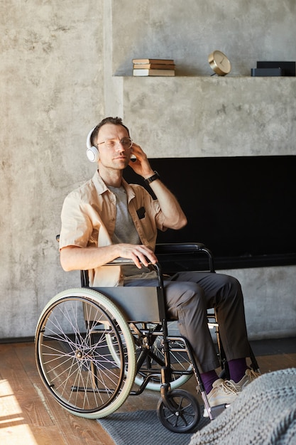 Vertikales Porträt in voller Länge eines behinderten erwachsenen Mannes im Rollstuhl, der zu Hause Musik hört