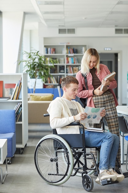 Vertikales Porträt in voller Länge eines fröhlichen behinderten Studenten, der mit der jungen Frau in der Hochschulbibliothek spricht