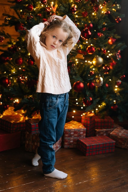 Vertikales Porträt in voller Länge eines hübschen kleinen blonden lockigen Mädchens, das in die Kamera blickt, die neben geschmücktem Weihnachtsbaum und Geschenkboxen mit Geschenken steht