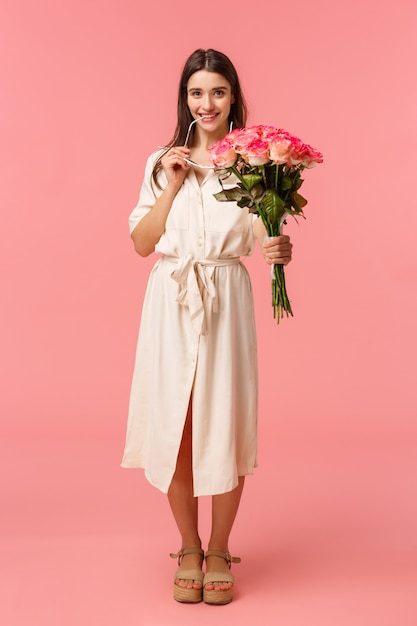 Vertikales Porträt in voller Länge sinnliche, romantische und kokette junge Frau, die etwas Interessantes sieht, mit Versuchung oder Verlangen schaut, Blumenstraußblumen hält, Rosen erhält, rosa Wand