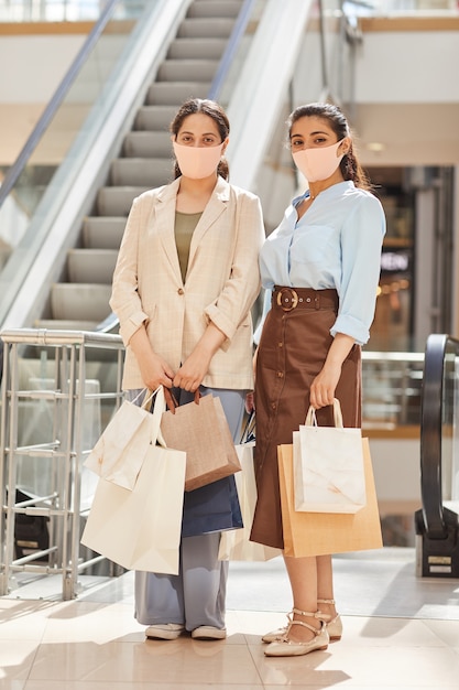 Vertikales Porträt in voller Länge von zwei jungen Frauen, die Masken beim Einkaufen im Einkaufszentrum und tragen