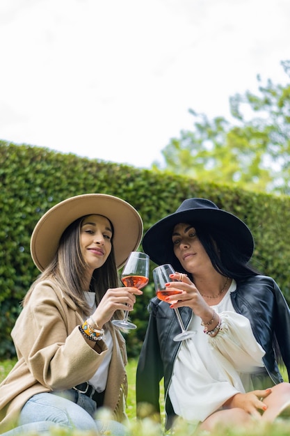 Vertikales Porträt schöner lächelnder Frauen mit Hüten, die im Park Wein trinken und anstoßen