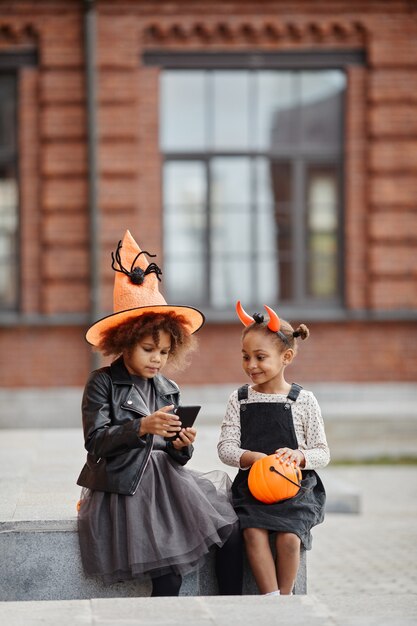 Vertikales porträt von zwei afroamerikanischen mädchen, die im freien halloween-kostüme tragen und smartph...