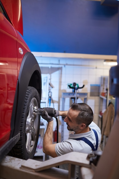 Vertikales Seitenansichtporträt des reifen Automechanikers, der Reifen auf rotem Auto in Autoreparaturwerkstatt, Kopienraum ändert