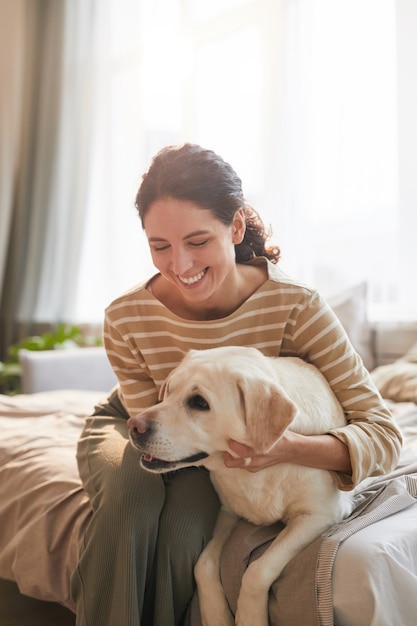 Vertikales, warm getöntes Porträt einer lächelnden jungen Frau, die den Hund umarmt, der auf dem Bett in einem gemütlichen Wohnhaus liegt, das von Sonnenlicht beleuchtet wird