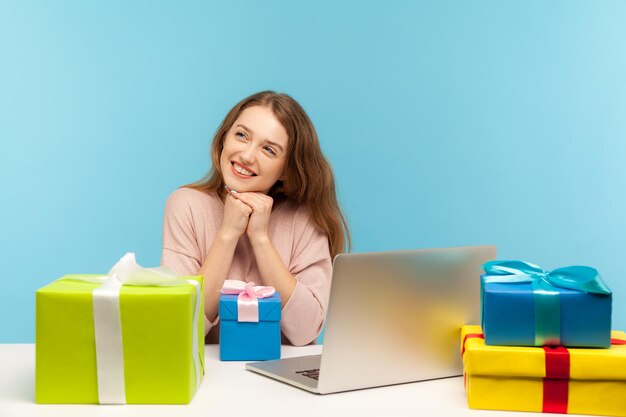 Verträumte Büroangestellte, die am Arbeitsplatz sitzt, freundlich lächelt, umgeben von vielen Geschenkboxen und angenehme Erinnerungen denkt, Geburtstag feiert, Berufsurlaub. Innenaufnahme, Studioaufnahme