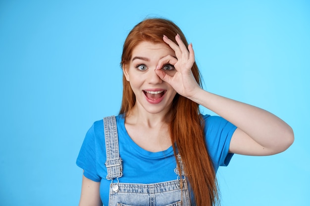 Verträumte, neugierige, attraktive, fröhliche Rothaarige Frau überrascht, schaut fasziniert Kamerablick durch die Finger Okay-Zeichen, steh amüsiert, bewundere blauen Hintergrund, schau dir die Promo an