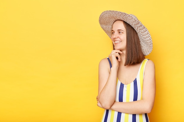 Verträumte, positive, optimistische Frau in gestreiftem Badeanzug und Hut isoliert auf gelbem Hintergrund, die lächelnd wegschaut und Platz für Werbung kopiert