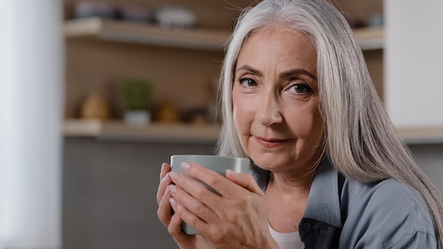 Verträumte reife Frau, die eine Tasse heißes Getränk hält und die Hände auf dem Becher wärmt, um eine Grippe zu verhindern, die wegschaut