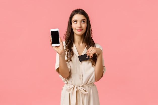 Verträumte und kokette gut aussehende junge Frau im Kleid, träumend davon, online auf Lieferauftrag zu warten, Smartphone und Kreditkarte zu halten, zeigt mobilen Bildschirm