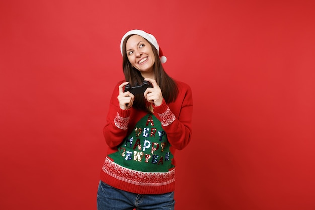 Verträumtes junges Santa-Mädchen in Pullover-Weihnachtsmütze, das aufschaut und Videospiel mit Joystick spielt, der auf rotem Wandhintergrund isoliert ist. Frohes neues Jahr 2019 Feier Urlaub Party Konzept. Kopieren Sie Platz.