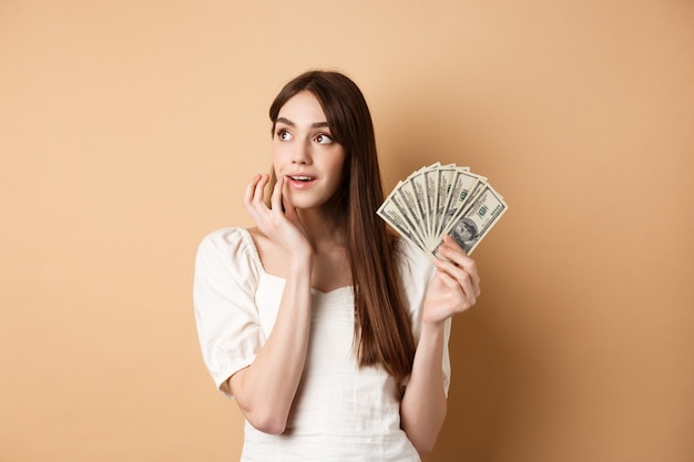 Verträumtes Mädchen, das Dollarnoten hält und über das Einkaufen nachdenkt, nachdenklich in die obere linke Ecke schaut, plant, wie man Geld verschwendet, auf beigem Hintergrund stehend.