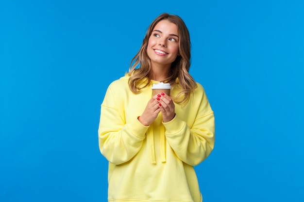 Verträumtes niedliches blondes europäisches Mädchen im gelben Kapuzenpulli, das romantisch und nachdenklich aufblickt und zu sich selbst lächelt, als sie etwas kawaii darstellt, das Pappbecher hält, der Kaffee trinkt