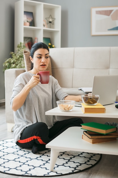 Verwirrt junges Mädchen benutzte Laptop mit einer Tasse Tee auf dem Boden hinter dem Couchtisch im Wohnzimmer